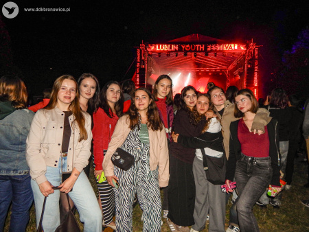 Noc. Koncert plenerowy. Grupa dziewczyn pozuje do zdjęcia. Za nimi widać scenę podświetloną na czerwono. Na niej napis: Lublin Youth Festival.