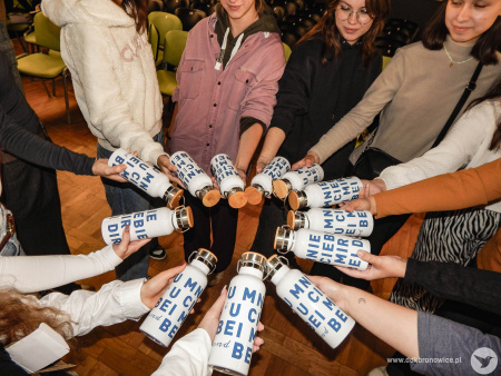 Grupa stoi w kole. Każdy z uczestników trzyma białą butelkę z niebieskim napisem.
