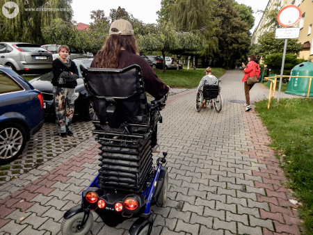 Dwie kobiety jadą na wózkach inwalidzkich. Obok nich Idą dwie kobiety. Jedna ubrana w czerwoną kurtkę. Druga ubrana w czarną bluzę i kolorowe dżinsy.
