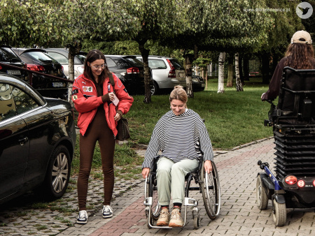 Osoba niepełnosprawna jeździ na wózku inwalidzkim. Jest uśmiechnięta. Kobieta ubrana jest czarną koszulkę w białe paski. Obok niej idzie kobieta w czerwonej kurtce. Obie kobiety uśmiechają się.
