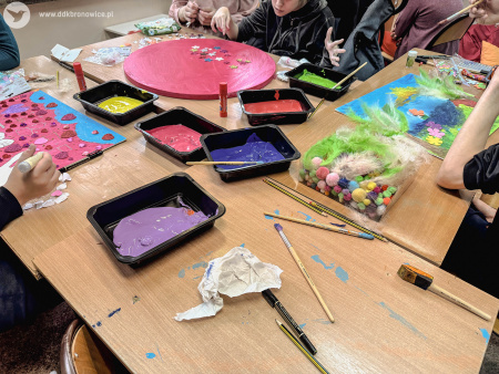 Stół warsztatowy. Na nim pędzle, farby i artykuły papiernicze. Widać pracujących przy stole uczestników wymiany.