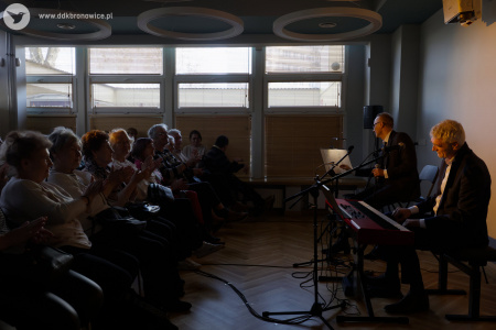 Po lewej stronie publiczność. Po prawej stronie artyści w czarnych garniturach. W tle duże okna.