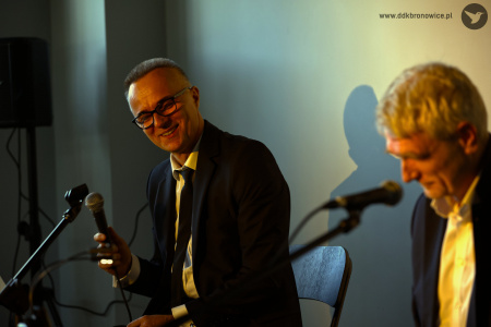 Artyści siedzą. Oświetleni są żółtym światłem. Patrycjusz Sokołowski po lewej stronie trzyma mikrofon.