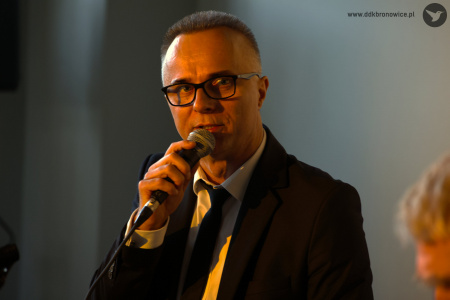 Patrycjusz Sokołowski. Trzyma blisko ust mikrofon. Oświetlony jest ciepłym, żółtym światłem. Patrzy w kierunku pianisty.