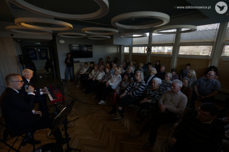 Po lewej stronie występują artyści. Po prawej stronie widać zgromadzoną widownię. W tle wiszący czarny telewizor. Na suficie okrągłe lampy.