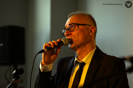 Patrycjusz Sokołowski śpiewa do mikrofonu emocjonalną piosenkę. Ubrany w czarny garnitur i czarny krawat. Oświetlony jest żółtym światłem.