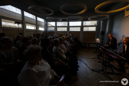 Po lewej stronie publiczność. Po prawej artyści oświetleni żółtym i ciepłym światłem. Przez okna pada zimne światło. Widać kontury osób na widowni.