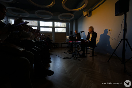 Artyści oświetleni żółtym światłem wykonują piosenkę. Po lewej stronie siedzi widownia.