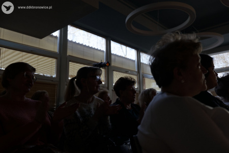 Osoby z widowni siedzą i słuchają koncertu. W tle światło pada przez okna.