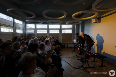 Artyści po prawej stronie kłaniają się publiczność. Widownia klaszcze w dłonie. Artystów oświetla delikatne żółte światło.