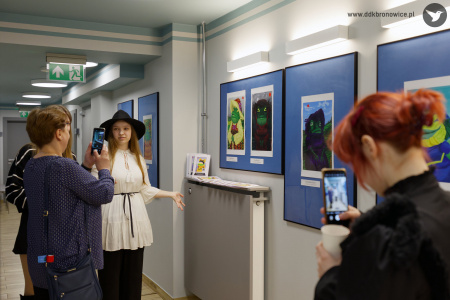 Kobieta w niebieskiej koszuli robi zdjęcie telefonem autorce prac. Autorka stoi na tle obrazów w niebieskich antyramach.