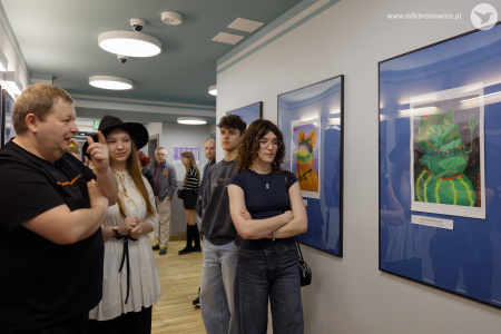 Publiczność ogląda wystawę. Po prawej stronie wzdłuż ściany z obrazami stoi mężczyzna i kobieta. Po lewej stronie autorka obrazów rozmawia ze starszym mężczyzną.