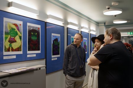 Dwóch mężczyzn i Oleksandra Khodakivska oglądają obrazy w niebieskiej antyramie i przedstawiają żaby w ubraniach. Jedna ma żółtą koszulę i kapelusz, a druga jest czarna.