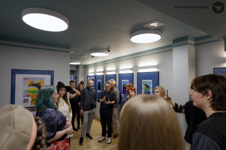 Osoby zgromadzone na wernisażu słuchają przemówienia Oleksandry Khodakivskiej. W tle na białej ścianie widać obrazy w niebieskich antyramach.