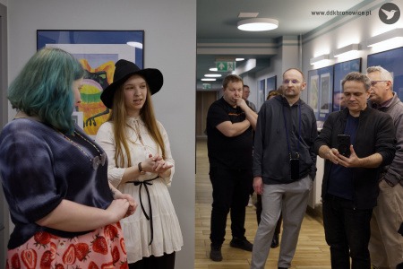 Autorka prac przemawia do zgromadzonych osób. Obok niej po lewej stronie stoi koordynatorka Galerii Przechodniej, Emilia Skubis. Po prawej stronie uczestnicy wystawy słuchają autorki.