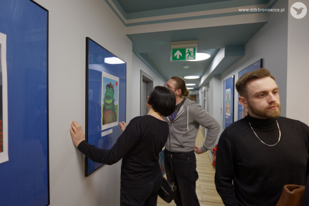 Kobieta w czarnej bluzce i mężczyzna w szarej bluzie oglądają obraz przedstawiający żabę. Żaba ma na sobie zielone ubranie. Po prawej stronie widać mężczyznę w czarnym golfie.