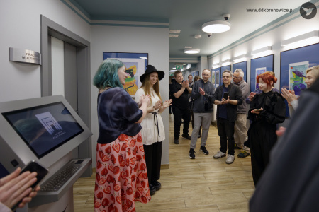 Publiczność zgromadzona na wystawie bije brawo. Autorka prac uśmiecha się. W tle w niebieskich antyramach wiszą obrazy.