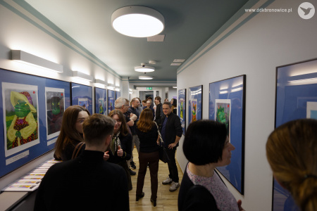 Galeria Przechodnia. Białe ściany. Na ścianach obrazy przedstawiające żaby w niebieskich antyramach. W korytarzu stoi grupa osób oglądających obrazy.