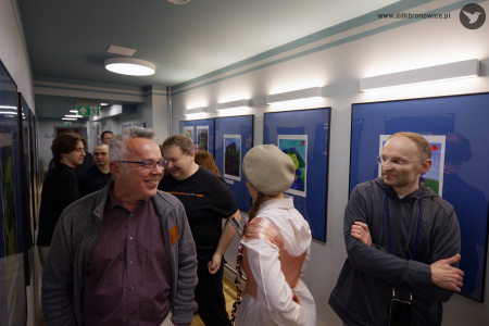 Dwóch mężczyzn uśmiecha się w korytarzy między zawieszonymi obrazami. Między nimi stoi kobieta w białej koszuli i beżowym berecie. W tle grupa osób.