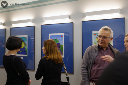 Po lewej stronie stoją dwie rozmawiające kobiety. Po prawej stoi mężczyzna w koszuli w kratę. Na ścianie zawieszone obrazy w niebieskich antyramach.