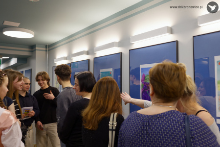 Na pierwszym planie rozmawiają trzy kobiety. Jedna z nich wskazuje palcem obraz. W tle grupa osób ogląda obrazy.