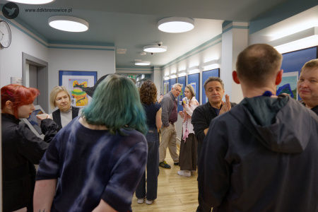 Grupy osób rozmawiają na wystawie. W tle widać kobietę w białej koszuli i beżowym berecie rozmawiającą w mężczyzną. Wskazuje ona na Emilię Skubis.