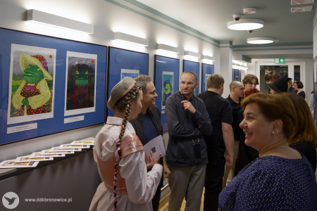 Kobieta w białej koszuli rozmawia z grupą uczestników wystawy. W tle wiszące obrazy przedstawiające kolorowe żaby w ubraniach.