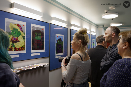 Kobieta w szarej bluzce ogląda obraz przedstawiające żabę. Żaba ubrana jest w beżową sukienkę i czerwony kapelusz.