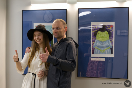 Mężczyzna w szarej bluzie pozuje do zdjęcie z Oleksandrą Khodakivską. W tle obrazy żab. Osoby pokazują kciuki.