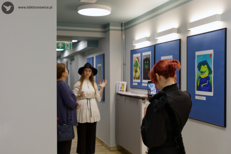 Kobieta w czarnej koszuli nagrywa autorkę prac. Autorka opowiada i wskazuje na swoje obrazy. Obok niej stoi kobieta w niebieskiej koszuli.