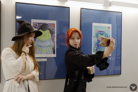 Kobieta w czarnej koszuli robi sobie zdjęcie telefonem na tle obrazów. Obok niej stoi autorka prac.
