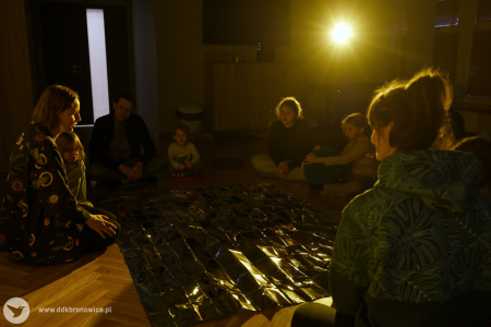 Grupa osób siedzi w kręgu. Między nimi rozłożony czarny, lśniący materiał. Na materiał i uczestników pada żółte światło.