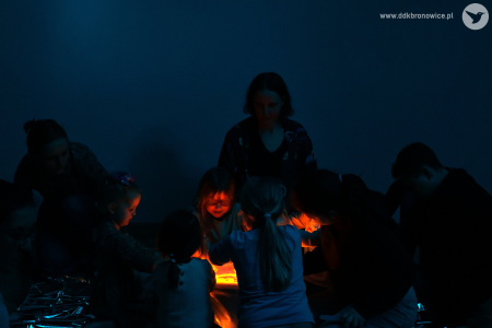 Ciemne pomieszczenie. Z pudełka wychodzi czerwone światło. Światło oświetla dzieci, które siedzą wokół.