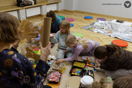 W pomieszczeniu zapalone światło. Czwórka dzieci tworzą kolorowe ozdoby na podłodze. Obok nich leżą nożyczki, papier i artykuły papiernicze.