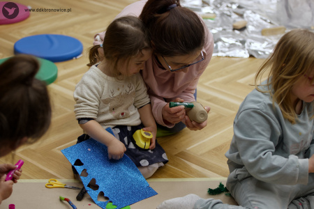Mama pomaga córce w tworzeniu kolorowej ozdoby. Dziewczynka trzyma niebieską kartkę. Widać na kartce wycięte serduszka.