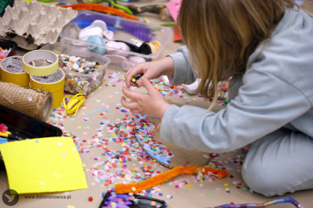 Dziewczynka w błękitnej bluzie tworzy kolorową ozdobę. Przed sobą na podłodze ma rozłożone małe, kolorowe kółka.