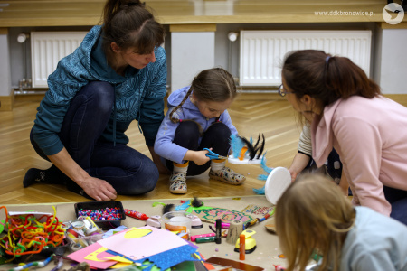 Mamy pomagają swoim córkom w tworzeniu ozdób. Siedzą na podłodze. Między nimi rozłożone nożyczki, taśmy, małe ozdoby i artykuły papiernicze.
