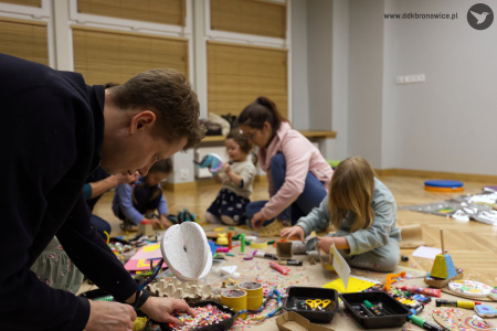 Mężczyzna pomaga w tworzeniu ozdoby. Nosi marynarkę. Naprzeciwko dziewczynka w błękitnym dresie tworzy ozdobę.