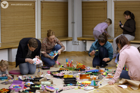 Uczestnicy na podłodze tworzą kolorowe ozdoby. W tle, dwójka dzieci na parapecie używa gorącego kleju.