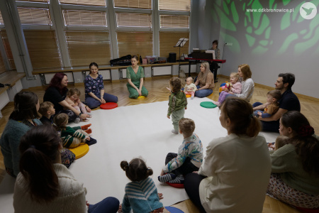 Pracownie Kultury Dziesiąta. Na białym dywanie siedzą uczestnicy wydarzenia. Małe dzieci siedzą na kolanach rodziców.