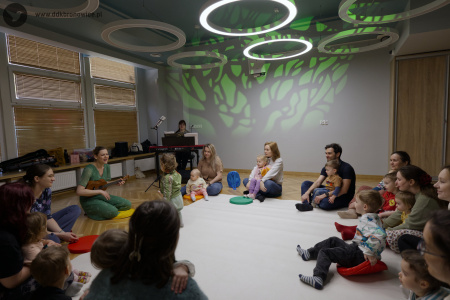 Uczestnicy siedzą na dywanie. W tle wyświetlone czarno-zielone drzewo. W tle po lewej stronie kobieta gra na czerwonym pianinie.