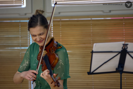 Prowadząca spotkanie, Sylwia Namroży, gra na skrzypcach w zielonej sukience. Bardzo szeroko się uśmiecha.Po prawej stronie pulpit z białymi kartkami.