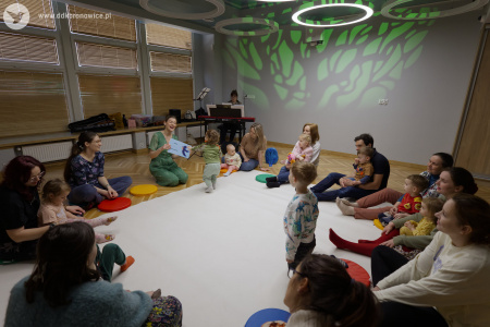 Uczestnicy bawią się na białym dywanie. W tle wyświetlone drzewo. W tle po lewej stronie kobieta grająca na pianinie.