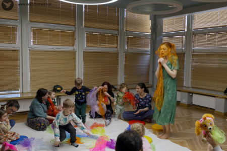 Prowadząca stoi. Na głowie ma złotą, przezroczystą chustę. Uczestnicy siedzą. Na białym dywanie rozłożone kolorowe i przezroczyste chusty.