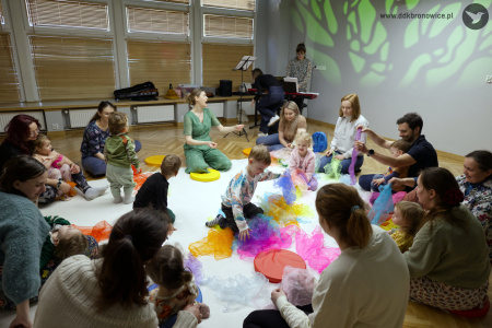 Dzieci bawią się kolorowymi chustami na białym dywanie. Prowadząca zajęcia siedzi razem z uczestnikami. Śpiewa.