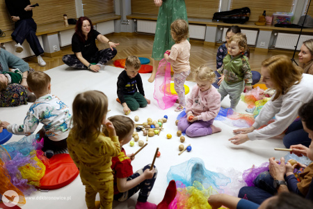 Na białym dywanie dzieci bawią się kolorowymi chustami. Na dywanie rozłożone są kulki. Rodzice bawią się ze swoimi dziećmi.