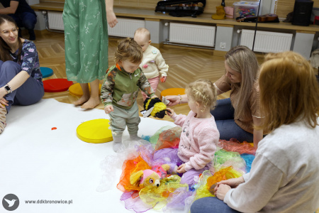 Dziewczynka w różowym swetrze siedzi na kolorowych chustach. Obok niej stoi mały chłopiec oboje trzymają pluszową pszczołę.
