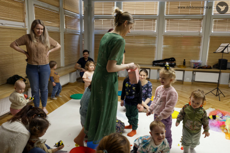 Sylwia Namroży stoi na środku białego dywanu. Wokół niej stoją uśmiechnięte dzieci. Prowadząca rozdaje z okolicznościowej torebki niespodzianki.