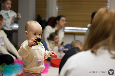 Małe dziecko ubrane w jasny sweterek trzyma pluszową pszczołę. Pokazuje pluszaka osobie dorosłej.