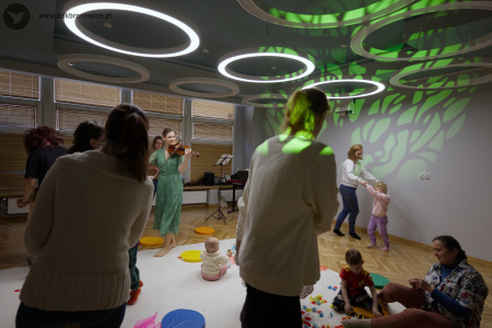 Prowadząca wydarzenie gra na skrzypcach. Po prawej stronie wyświetlone zielone drzewo. Rodzice tańczą ze swoimi dziećmi.